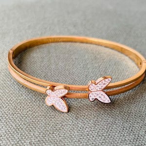 May include: Gold-coloured bangle bracelet with two butterfly features. Each butterfly is decorated with small, sparkling crystals. The bracelet has a double-band design and is shown on a textured, grey surface. A piece of jewellery.