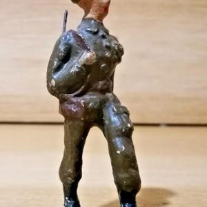 May include: A vintage metal toy soldier figurine standing on a green base. The figure wears a dark olive green uniform with a matching helmet, a brown satchel, and carries a rifle on the back. The paint shows signs of wear and age throughout.