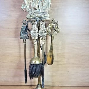 Puede incluir: Un juego de herramientas para chimenea de latón con un soporte coronado por un emblema de barco. El set incluye un atizador, un cepillo con cerdas negras, una pala y un calzador, cada uno con mangos decorativos que representan barcos y una cabeza de caballo.