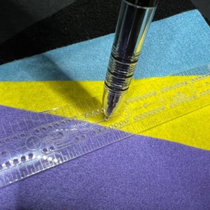 May include: A clear plastic ruler with a grid pattern and holes is placed on top of layers of purple, yellow, and blue felt. A silver pen is positioned on the ruler.