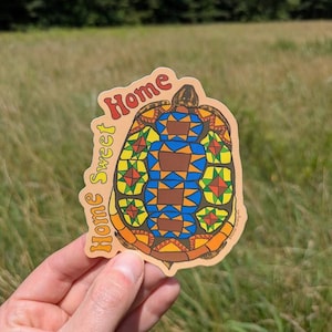 May include: A colorful sticker featuring a turtle design with a geometric pattern in shades of blue, orange, yellow, and green. The sticker has the words "Home Sweet Home" in a retro font.
