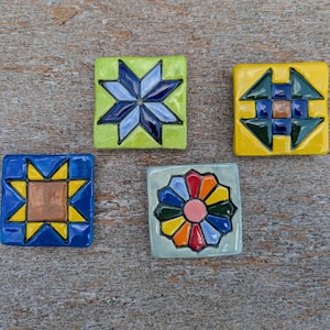 May include: Five colorful, square ceramic tiles with geometric designs. The tiles feature a variety of patterns, including stars and floral motifs, in shades of blue, yellow, green, orange, and red. The tiles are arranged on a wooden surface.
