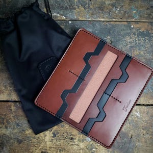 Peut inclure: Un portefeuille en cuir marron avec des accents géométriques noirs et une bande centrale beige. Le portefeuille est à côté d'un sac en tissu noir. Le mot "Farlee" est estampé sur le portefeuille.