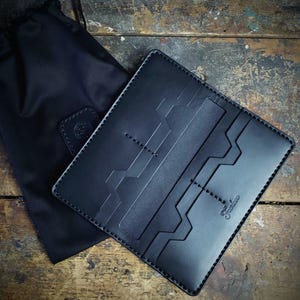 May include: Black leather wallet with a geometric design and stitched detailing. The wallet is open, showing multiple card slots. A black drawstring bag is next to the wallet. The word "Santino" is embossed on the wallet.