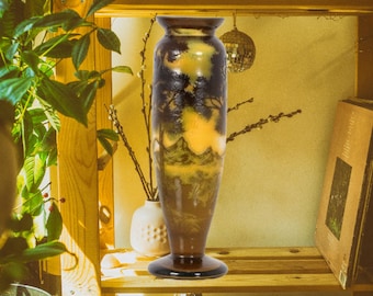 May include: A brown and yellow ceramic vase with a landscape scene painted on it. The vase has a wide base and a narrow neck. It is sitting on a wooden shelf.