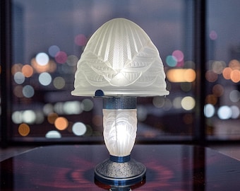 May include: A decorative table lamp with a frosted glass shade and base. The shade has a detailed, textured design, and the base is a dark blue color. The lamp is illuminated, casting a soft glow. The background features blurred city lights.