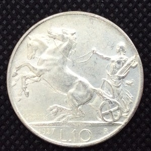 May include: A silver Italian coin with a depiction of a chariot pulled by two horses. The coin has the year 1927 and the denomination L.10 on it.