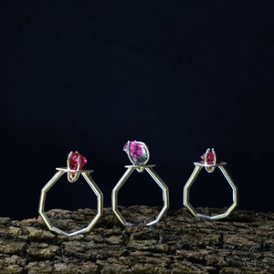 Op de afbeelding: Drie zilveren ringen met achthoekige banden en rode edelstenen in het midden. De ringen worden getoond op een stuk verweerd hout.