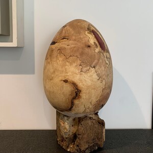 May include: A large, wooden egg sculpture with a natural wood finish and a reddish-brown resin inlay. The egg is sitting on a wooden base.