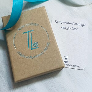 Puede incluir: Una caja de joyer&iacute;a marr&oacute;n con el texto "Tressia Lily Designs" y "Sterling Silver Handmade Jewellery" en color turquesa. Una cinta turquesa est&aacute; encima de la caja. Una tarjeta blanca con el texto "Your personal message can go here" est&aacute; al lado de la caja.