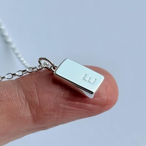 May include: A silver pendant necklace with a rectangular charm, engraved with the letter "E". The charm hangs from a fine chain. The necklace is displayed against a light background.