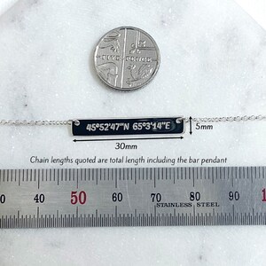 Puede incluir: Un collar con colgante de barra plateada grabado con coordenadas, que mide 30 mm de largo y 5 mm de ancho. El collar se muestra con una moneda y una regla, que indica la longitud de la cadena. El texto en el colgante dice "45&deg;52'47"N 65&deg;3'14"E".