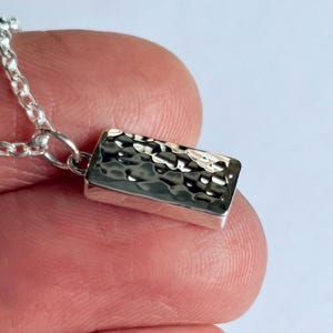 May include: A silver rectangular pendant with a hammered texture, hanging from a delicate chain. The pendant is a small, rectangular bar with a textured surface, reflecting light. The chain is also silver.