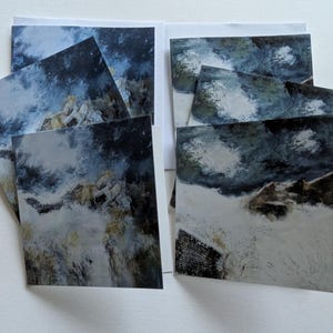 Puede incluir: Un conjunto de impresiones artísticas cuadradas que presentan una pintura de paisaje abstracto. La obra de arte utiliza una paleta de azules, grises y blancos para representar una escena montañosa con un cielo dramático. Las impresiones están dispuestas sobre una superficie blanca.