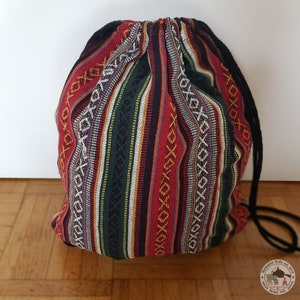 Könnte beinhalten: Ein Rucksack mit Kordelzug und einem lebendigen Streifenmuster in Rot, Schwarz, Grün, Gelb und Weiß. Die Tasche hat einen schwarzen Kordelzugverschluss und besteht aus einem gewebten Stoff. Die Tasche steht auf einem Holzboden.