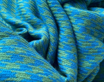 Può includere: Un primo piano di un tessuto intrecciato blu e verde con un motivo a chevron. Il tessuto è morbido e testurizzato, ed è piegato in modo da mettere in risalto il motivo.