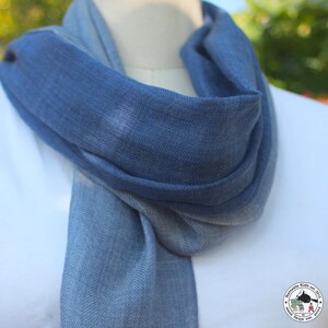 May include: A blue and gray scarf with a herringbone pattern is draped around a mannequin's neck. The scarf transitions from a darker blue to a lighter gray-blue. The scarf is made of a soft, woven material.