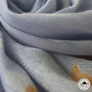 Könnte beinhalten: Nahaufnahme eines weichen, hellblauen Schals mit strukturiertem Gewebe. Der Stoff ist in sanften Falten drapiert, wodurch Schatten und Lichteffekte entstehen. Der Schal hat ein subtiles, fast aquarellartiges Aussehen. Die Ecke des Schals hat ein Logo mit der Aufschrift "Namaste Kids est. 2015".