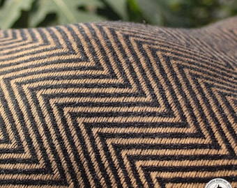 May include: Close-up of a textile with a repeating chevron pattern. The design features alternating black and tan zigzags, creating a textured surface. The fabric appears to be woven, with visible threads. The background is blurred green foliage.