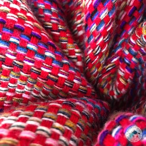 Puede incluir: Primer plano de un tejido tejido con una base roja vibrante. La tela presenta un patrón de tejido de cesta con hilos blancos, azules, morados y amarillos. La textura parece suave y ligeramente texturizada, con detalles de hilo visibles.