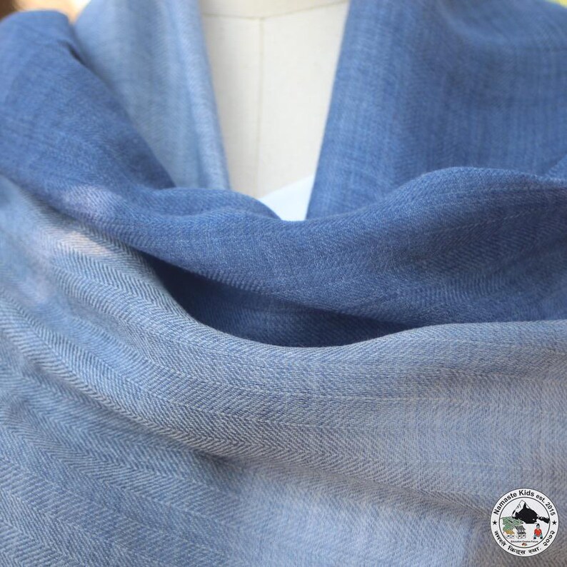 May include: A soft, blue scarf with a subtle herringbone pattern. The scarf transitions from a lighter shade of blue to a deeper, richer hue. The fabric appears to be lightweight and drapes elegantly, suitable for various styles and occasions.