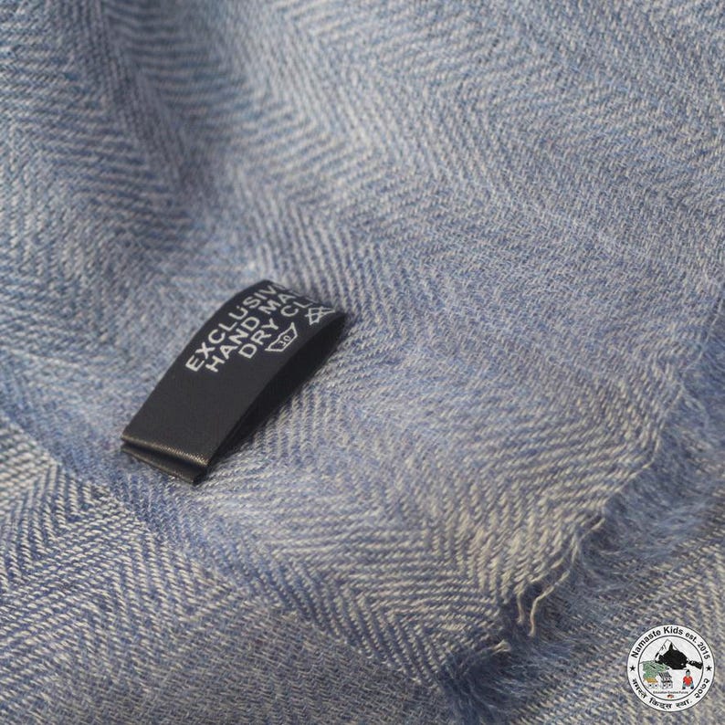 May include: Close-up of a light blue fabric with a herringbone pattern. A black tag with white text that reads "EXCLUSIVE HAND MADE DRY CLEAN" is attached to the fabric. The fabric has a soft, textured appearance.
