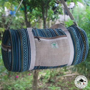 May include: A cylindrical duffel bag with a blue, green, and yellow striped pattern. The bag has a tan canvas pocket and straps, with a small green and white label that reads "Himalayan Hemp The Free". The bag is hanging from a tree.