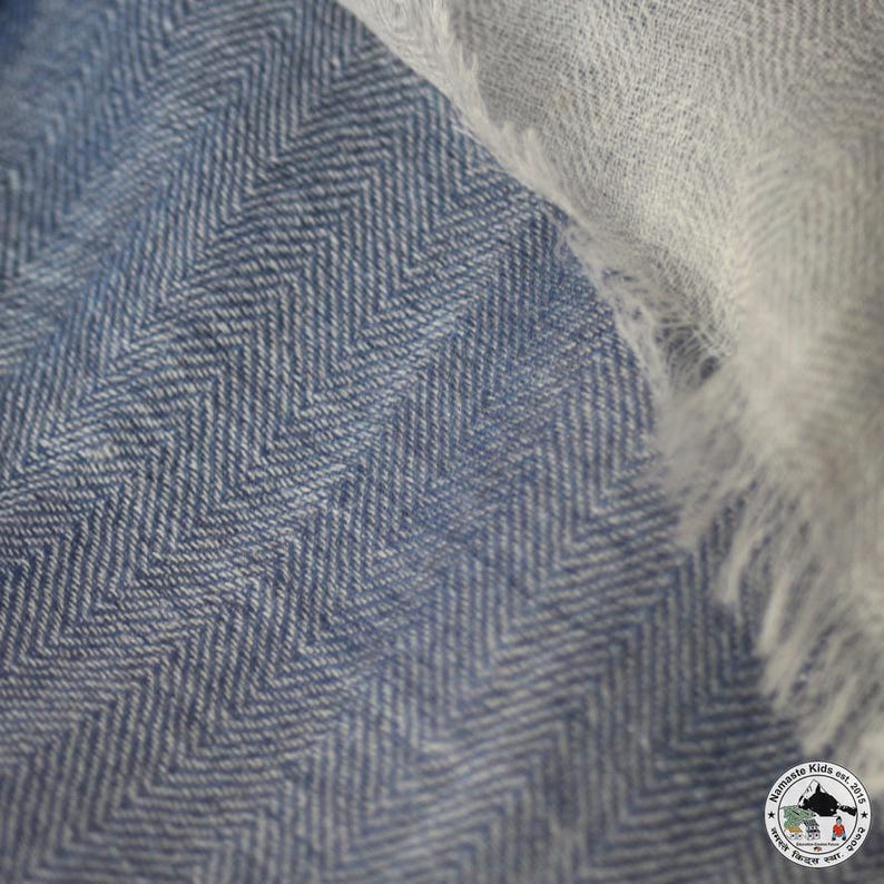 May include: Close-up of a blue and white herringbone patterned fabric. The fabric has a textured appearance with a diagonal weave. A section of white, fringed fabric is visible in the upper right corner, contrasting with the blue.