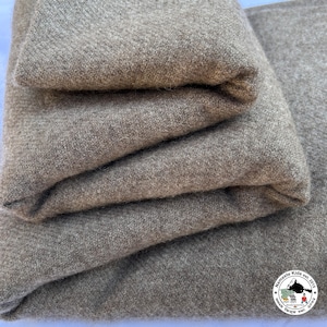 Puede incluir: Pila de tela beige doblada, posiblemente lana o una mezcla de lana. El material parece grueso y tejido. Los bordes están cuidadosamente doblados, lo que sugiere una manta o un producto textil. Un logotipo es visible en la esquina inferior derecha.