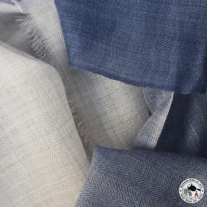 May include: Close-up of soft, textured fabric in shades of blue and off-white. The fabric has a subtle herringbone pattern. The image shows the texture and drape of the material, suggesting it could be a scarf or shawl.