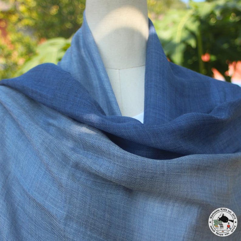 May include: A blue and gray scarf draped over a mannequin. The scarf has a gradient effect, transitioning from a deep blue to a lighter gray-blue. The fabric appears soft and woven, with a subtle texture. The scarf is likely made of a lightweight material.