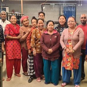 May include: A group of people, both men and women, stand together in a workshop setting. They are dressed in casual clothing, and some are smiling at the camera. The background includes industrial equipment and a high ceiling.