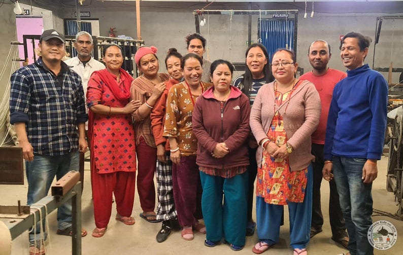 May include: A group of people, both men and women, stand together in a workshop setting. They are dressed in casual clothing, and some are smiling at the camera. The background includes industrial equipment and a high ceiling.