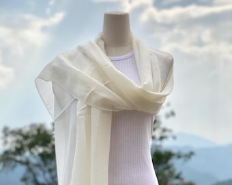 May include: A cream-colored scarf draped over a white ribbed tank top on a mannequin. The scarf is lightweight and appears to be made of a sheer fabric. The background features a cloudy sky and a mountain range.