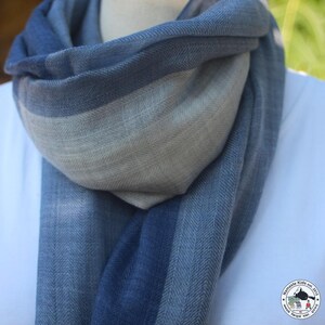 May include: A blue and gray scarf with a herringbone pattern. The scarf is draped around a white shirt. The scarf transitions from a deep blue to a lighter gray color. The scarf is made of a soft, woven material.