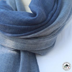 May include: A blue and gray scarf with a herringbone pattern. The scarf is made of soft fabric and has a gradient effect, transitioning from a deep blue to a lighter gray. The scarf is folded, showing the texture and layers.