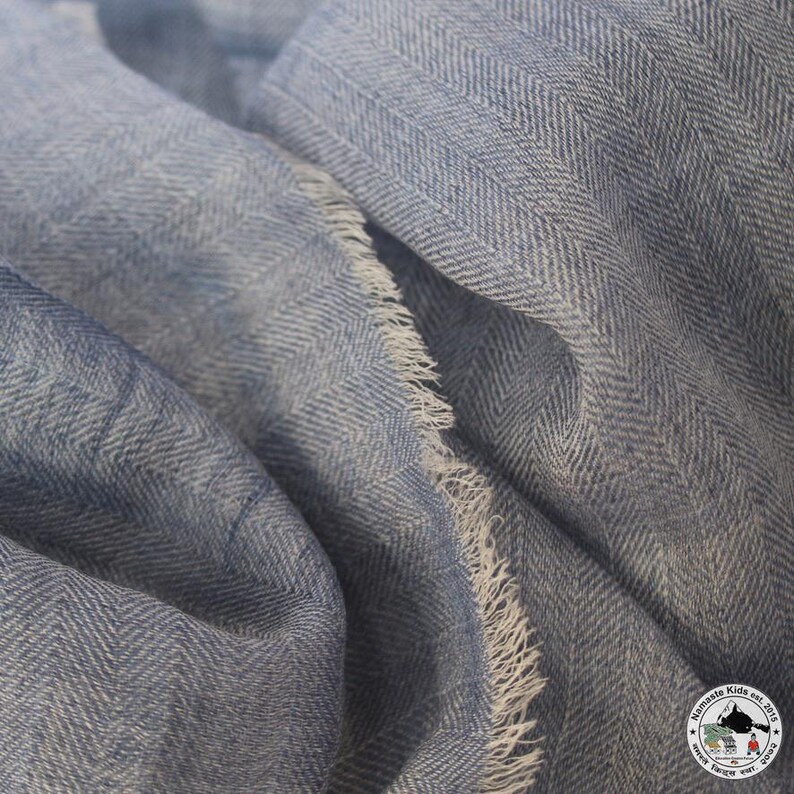 May include: Close-up of a light blue scarf with a herringbone pattern. The fabric has a soft texture and frayed edges. The scarf is folded, showing the intricate weave and color variations.