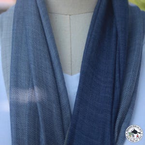 May include: A blue scarf with a herringbone pattern is draped over a mannequin. The scarf has a gradient effect, transitioning from a lighter blue to a darker shade. The fabric appears soft and lightweight, suitable for layering.