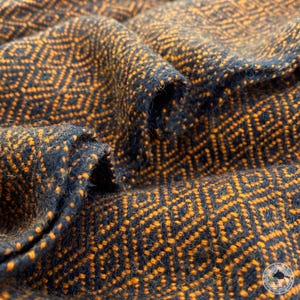 Op de afbeelding: Close-up van een zwarte en oranje geweven stof met een geometrisch patroon. De stof is zacht en textuurrijk, met een subtiele glans.