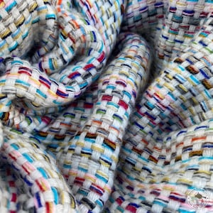 Può includere: Primo piano di un tessuto bianco tessuto con fili colorati intrecciati su tutta la stoffa. Il tessuto ha un aspetto testurizzato e tessuto a mano.