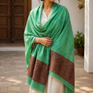 Può includere: Un grande scialle rettangolare di un vibrante colore verde menta con un sottile motivo tonale su tutta la superficie. Il bordo inferiore è rifinito con un'ampia fascia a contrasto marrone scuro, decorata con un intricato motivo paisley.