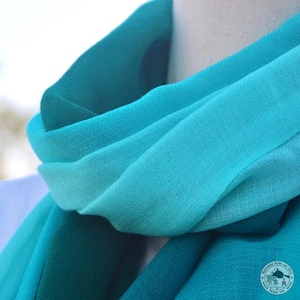 Op de afbeelding: Een zachte, turquoise blauwe ombre sjaal gemaakt van een lichtgewicht stof. De sjaal is gevouwen en gedrapeerd over de nek van een mannequin.