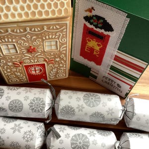 Può includere: Una scena festiva natalizia con un contenitore a forma di casa di pan di zenzero, un biglietto di Natale con un disegno di cassetta postale ricamato a punto croce e diversi cracker natalizi con motivi a fiocco di neve argentati e bianchi. Il biglietto dice "Buon Natale".