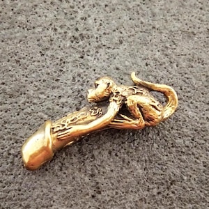 Può includere: Una scultura in metallo color oro di una scimmia che tiene in mano un oggetto fallico. La scimmia è dettagliata con pelliccia e lineamenti del viso. La scultura è su una superficie grigia strutturata.