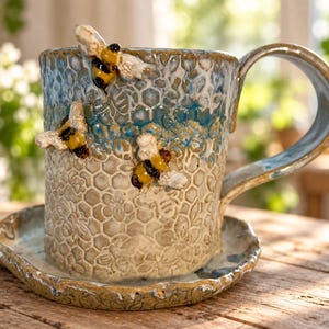 May include: A ceramic mug and matching saucer featuring a textured honeycomb and floral pattern. The mug is decorated with three sculpted, hand-painted bees in yellow, black, and white, with a blue and beige glaze finish and a curved handle.
