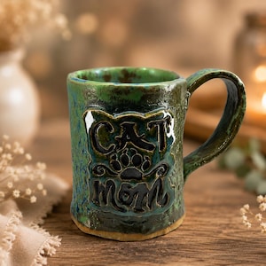 May include: A handmade ceramic mug with a textured green and blue glaze. The front features a raised, dark-colored relief design with the text CAT MOM and a central paw print icon. The mug has a sturdy handle and a cylindrical shape with a base.