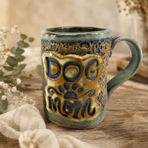 May include: A handmade ceramic mug with a blue and brown glaze, featuring a textured surface and a raised central panel with the embossed text DOG MOM and a paw print icon. The mug has a sturdy handle and a cylindrical shape with a rustic finish.