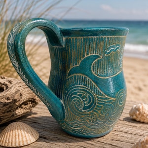 May include: A teal ceramic mug with a wave design, set against a beach backdrop. The mug features a detailed carving of ocean waves and a textured handle. Seashells are scattered around the mug on a wooden surface.