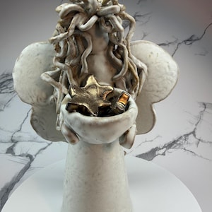 May include: A ceramic angel figurine with outstretched wings and curly hair. The angel holds a small bowl containing gold stars. The figurine is primarily white with brown accents and sits on a white base.