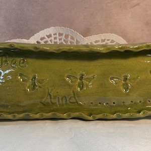 May include: A rectangular, handmade ceramic tray in a muted green hue. The tray features embossed bee designs and the words "Bee Kind." The edges have a wavy, textured design. The tray is placed on a white lace doily.